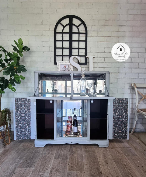 A gray cabinet with bottles inside is against a white brick wall. It has a decorative mirror on top, flanked by a potted plant. A semicircular window decor and a round sign are above. Flooring is wood-patterned.