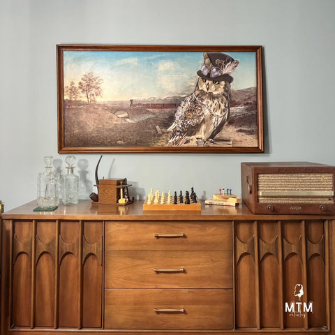 A mid-century modern wooden sideboard is adorned with various items: a set of crystal decanters, a small wooden box, a chess set, and a vintage radio. Above the sideboard, a framed painting features an owl wearing a hat and goggles in a scenic landscape.
