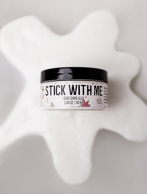 A round container labeled "Stick With Me Dixie Shine Glue 3.38 oz | 100 mL" sits on a white, glossy, spilled substance against a light background.