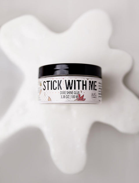 A round container labeled "Stick With Me Dixie Shine Glue 3.38 oz | 100 mL" sits on a white, glossy, spilled substance against a light background.