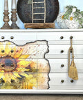 A beautifully refurbished painted dresser with a vibrant Sunny Daze A1 Rice Decoupage Paper illustration across its drawers, adorned by bronze knobs, gold tassels, and flanked by decorative home accessories.