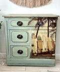 A vintage wooden cabinet with an ocean-themed design. The front features three drawers and a door with a beach scene of surfboards leaning against palm trees on it. The cabinet is painted light blue with distressed details. A potted plant is next to it.