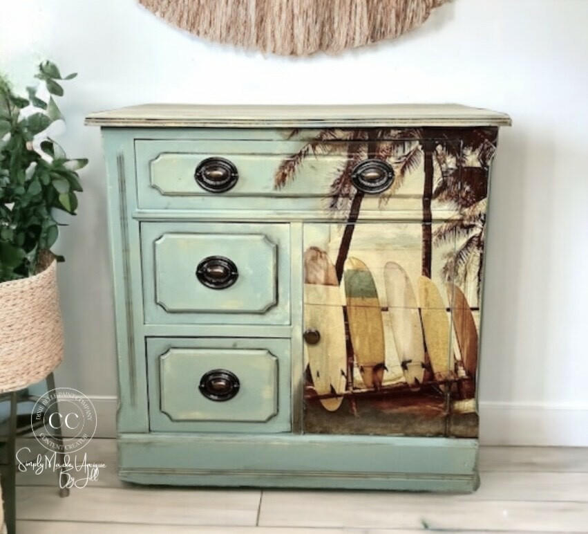 A vintage wooden cabinet with an ocean-themed design. The front features three drawers and a door with a beach scene of surfboards leaning against palm trees on it. The cabinet is painted light blue with distressed details. A potted plant is next to it.