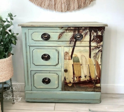A vintage wooden cabinet with an ocean-themed design. The front features three drawers and a door with a beach scene of surfboards leaning against palm trees on it. The cabinet is painted light blue with distressed details. A potted plant is next to it.