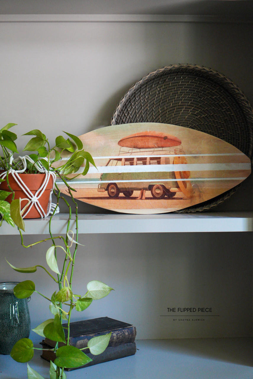 A decorative shelf displaying the Surf's Up Decoupage Paper on a small surfboard, accompanied by woven baskets, a potted plant in macrame, and a stack of books. The setting conveys a cozy, artistic vibe.