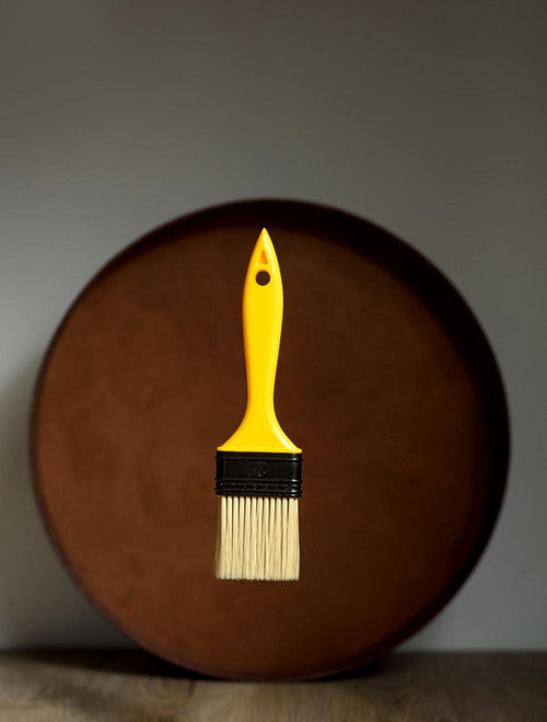 A yellow paintbrush with light bristles is suspended in front of a large, round, brown object against a plain background.