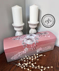 A beautifully painted pink chest adorned with floral designs is placed on a wooden table. Behind it, two white candles in ornate candle holders stand against a light-colored wall. A watermark on the wall reads "AJ's Vintage Designs." A decorative branch lies in the foreground.