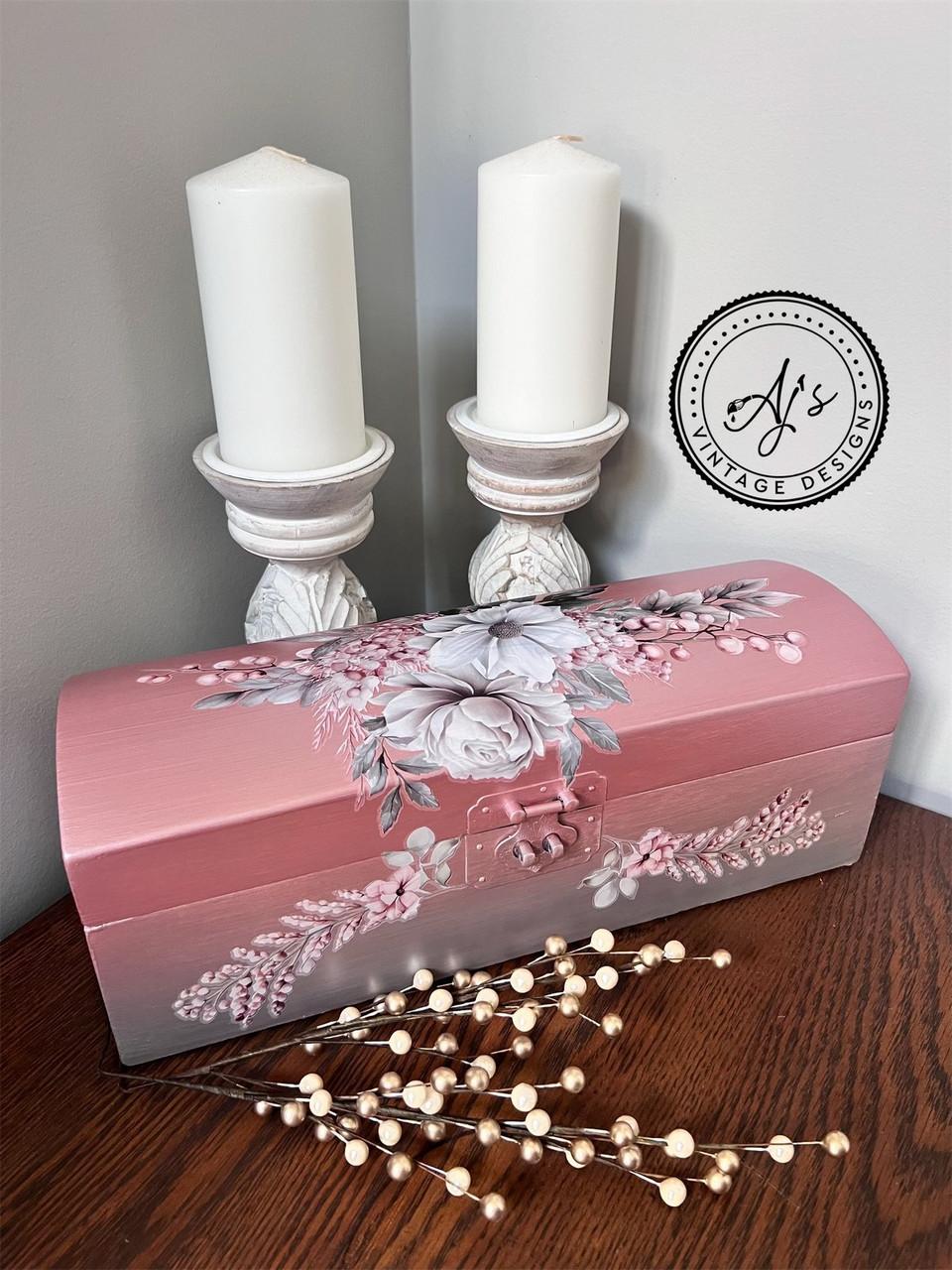 A beautifully painted pink chest adorned with floral designs is placed on a wooden table. Behind it, two white candles in ornate candle holders stand against a light-colored wall. A watermark on the wall reads "AJ's Vintage Designs." A decorative branch lies in the foreground.
