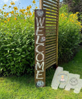 A wooden sign stands in a garden with yellow flowers. The sign vertically spells "WELCOME" in large white letters, accented with white floral designs. The background features lush greenery and a wooden slat structure. Two logos are visible in the bottom right corner.