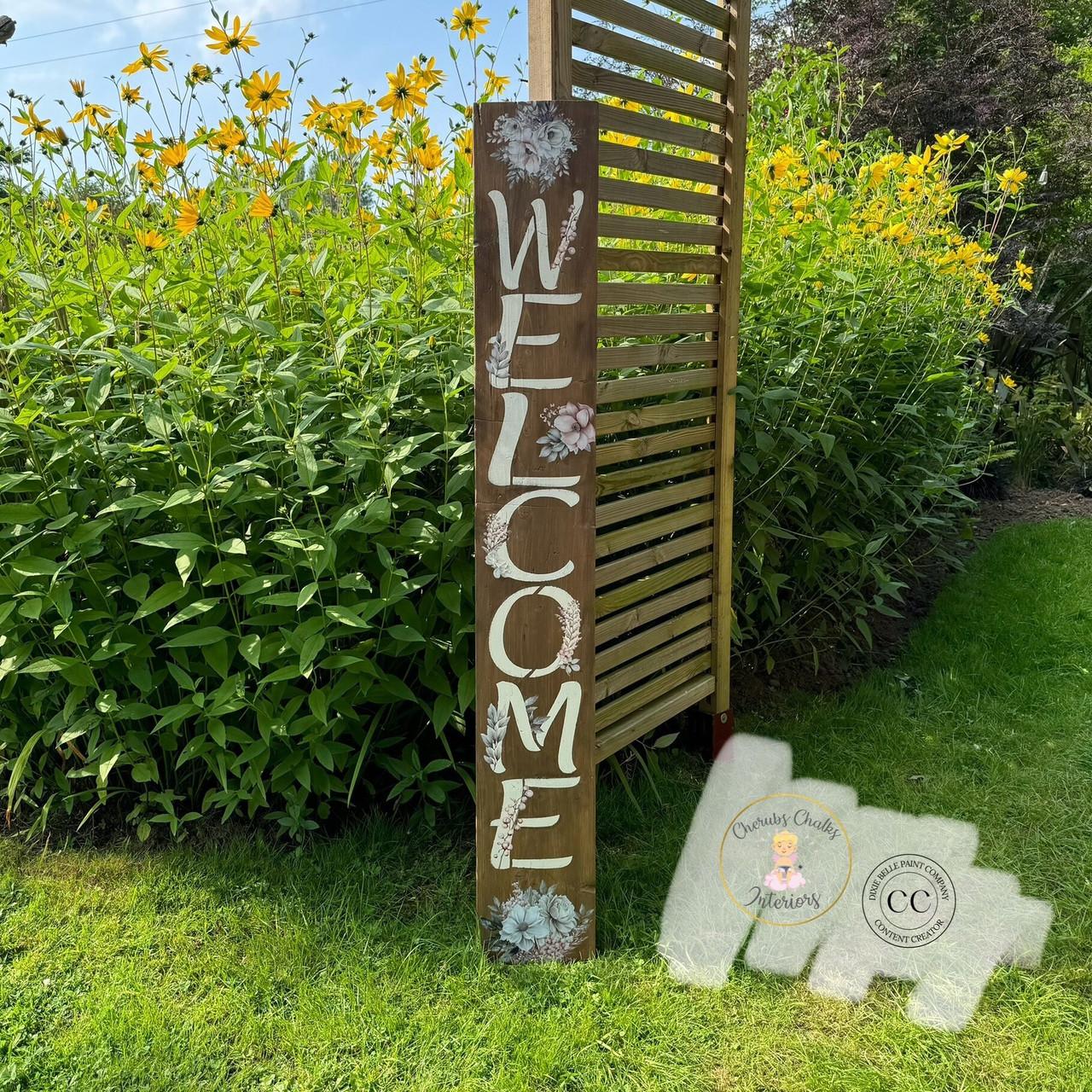 A wooden sign stands in a garden with yellow flowers. The sign vertically spells "WELCOME" in large white letters, accented with white floral designs. The background features lush greenery and a wooden slat structure. Two logos are visible in the bottom right corner.
