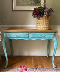Blue Agave Terra Clay Paint provides endless possibilities for your imagination and can be used on wood, canvas, tile, fabric and more! Entryway table with wood finish top painted with Blue Agave Terra Clay paint with distressed drawers. Staged in a room with wooden floor and tan wall with cut off picture frame. Adorned with a tall woven basket with faux flower pieces.