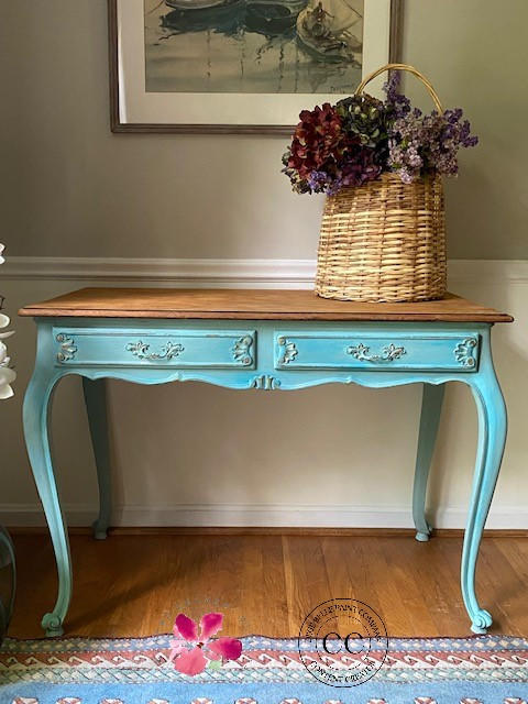 Blue Agave Terra Clay Paint provides endless possibilities for your imagination and can be used on wood, canvas, tile, fabric and more! Entryway table with wood finish top painted with Blue Agave Terra Clay paint with distressed drawers. Staged in a room with wooden floor and tan wall with cut off picture frame. Adorned with a tall woven basket with faux flower pieces.