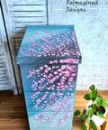 Blue Moon Terra Clay Paint provides endless possibilities for your imagination and can be used on wood, canvas, tile, fabric and more!
A beautiful cabinet using Blue Moon Terra Clay Paint as a base color with darker blues layered on top, with hand painted cherry bloosm branches. Staged next to small white planter with plant inside.
