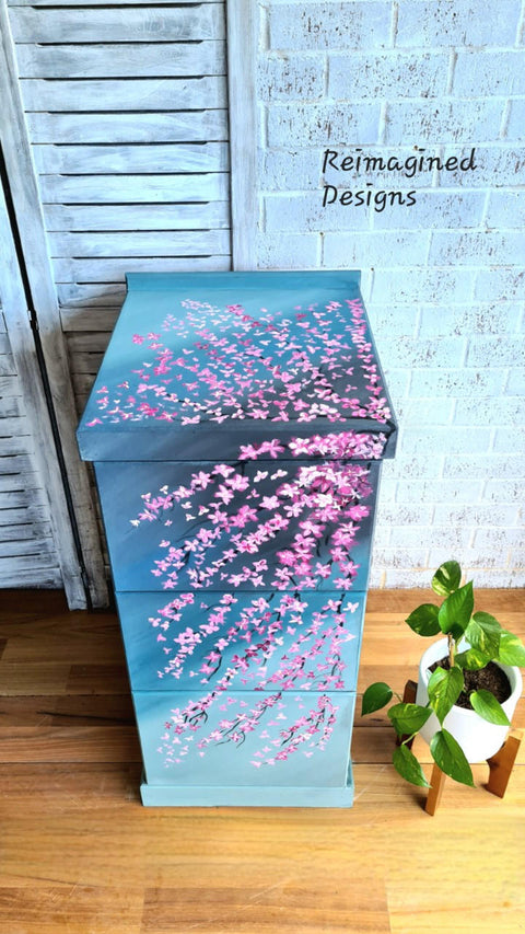 Blue Moon Terra Clay Paint provides endless possibilities for your imagination and can be used on wood, canvas, tile, fabric and more!
A beautiful cabinet using Blue Moon Terra Clay Paint as a base color with darker blues layered on top, with hand painted cherry bloosm branches. Staged next to small white planter with plant inside.