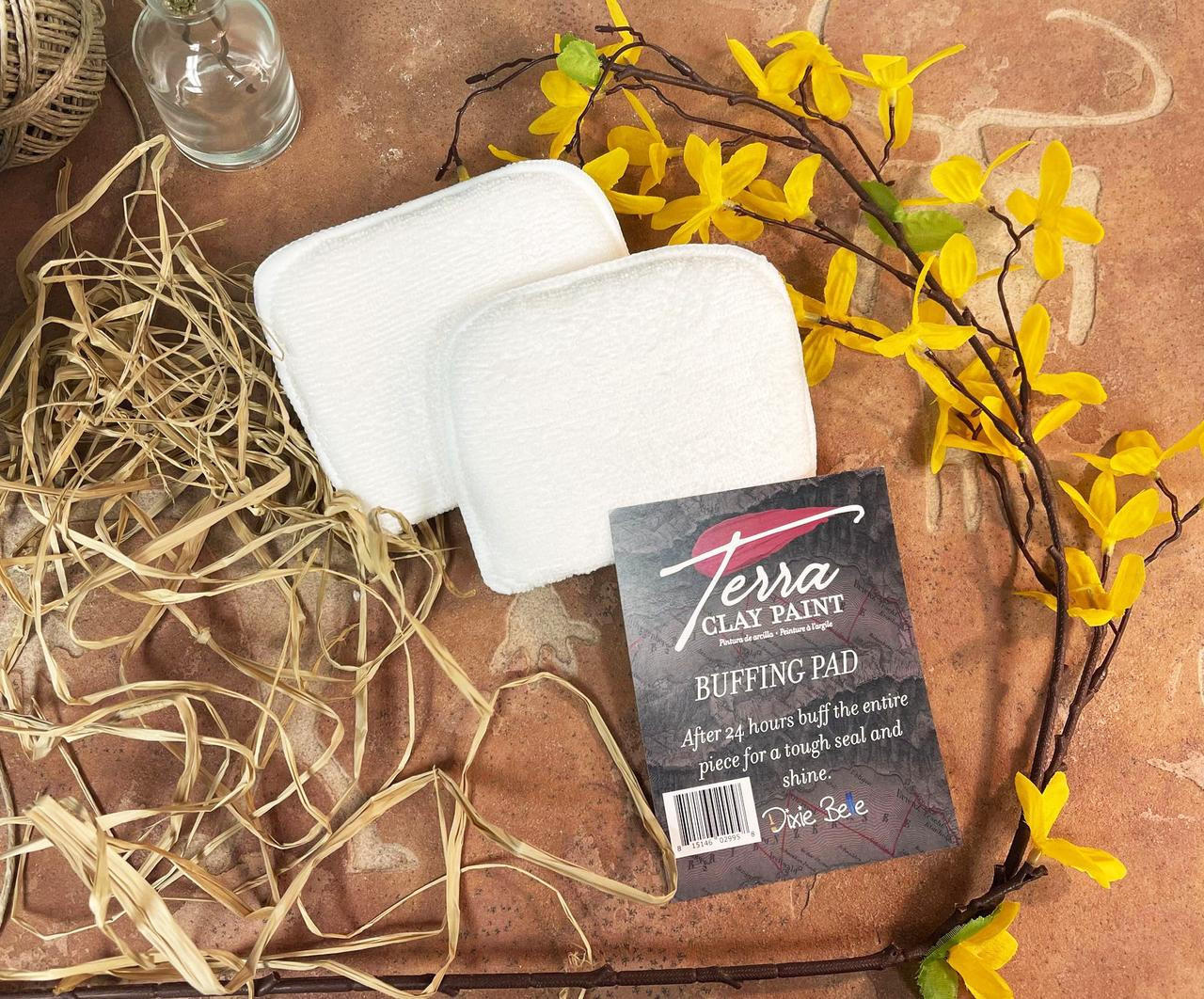 Terra Clay Paint Buffing Pad. Image of the Buffing Pad on top of a brown with animal carvings background. Staged with a branch with yellow flower and brown rope.