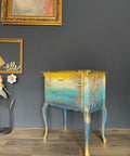 Daffodil Terra Clay Paint provides endless possibilities for your imagination and can be used on wood, canvas, tile, fabric and more! A side table that has a dark wooden top and has daffodil at the top of the base blended into white and then blue with gold accents.