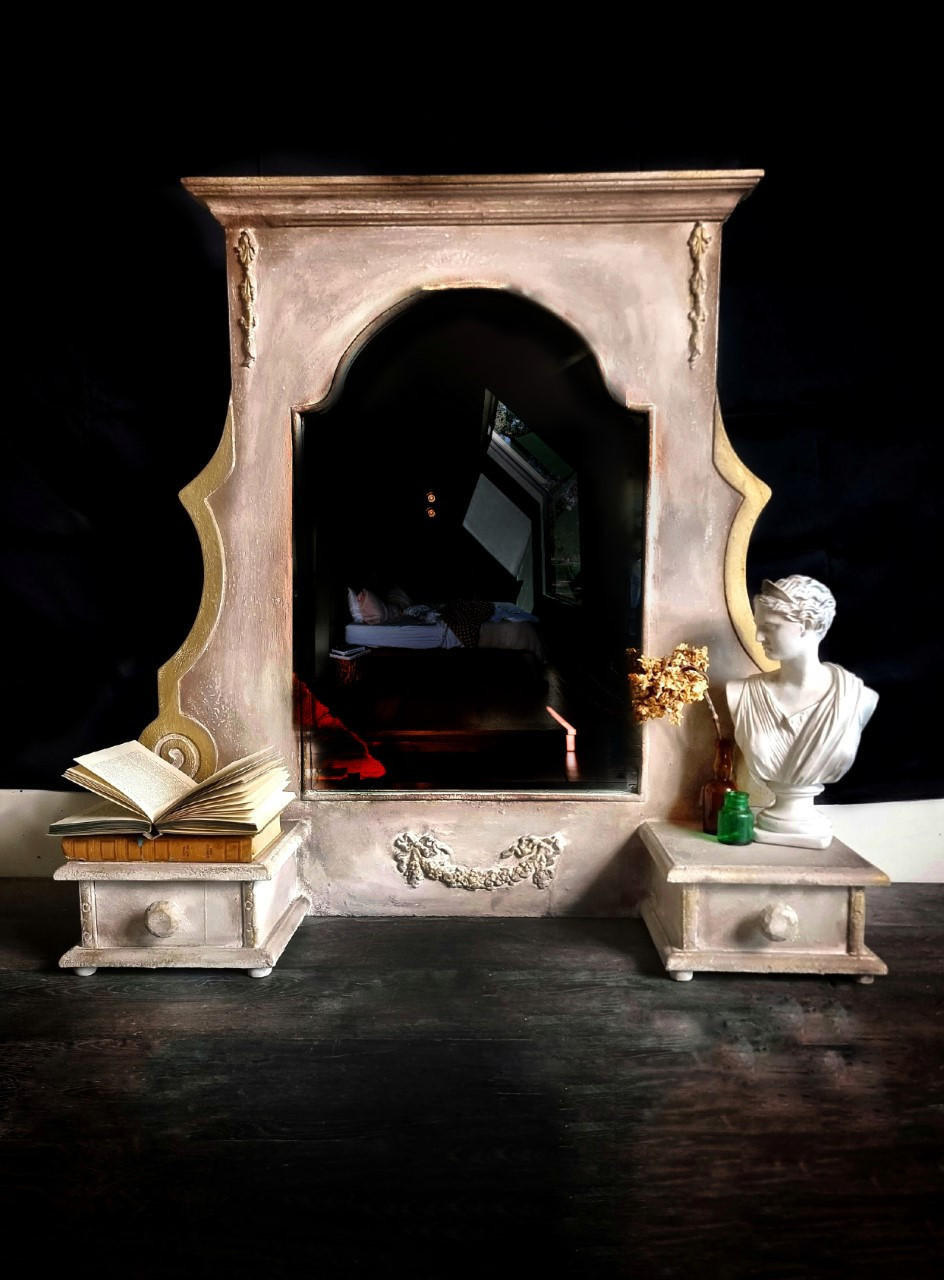 Desert Tan Terra Clay Paint provides endless possibilities for your imagination and can be used on wood, canvas, tile, fabric and more!
This is a stone surround for a  fireplace painted in Desert Tan Terra Paint.  There is an applique on the bottom of the piece.  It rests on a very dark cement floor and black background.  It is staged with a Greek Bust, dried flower in a green vase and a couple books, one which is splayed open.