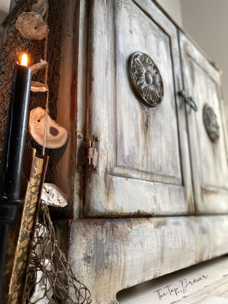 Terra Clay Paint Desert Tan Terra Clay Paint 
This is a very distressed wooden bench with two doors on the front.  There are appliques on both doors.  To the left is a lit, black candle, mesh, brown rope with oyster shells hanging off of it.  There is also a brown book and a branch on the floor.