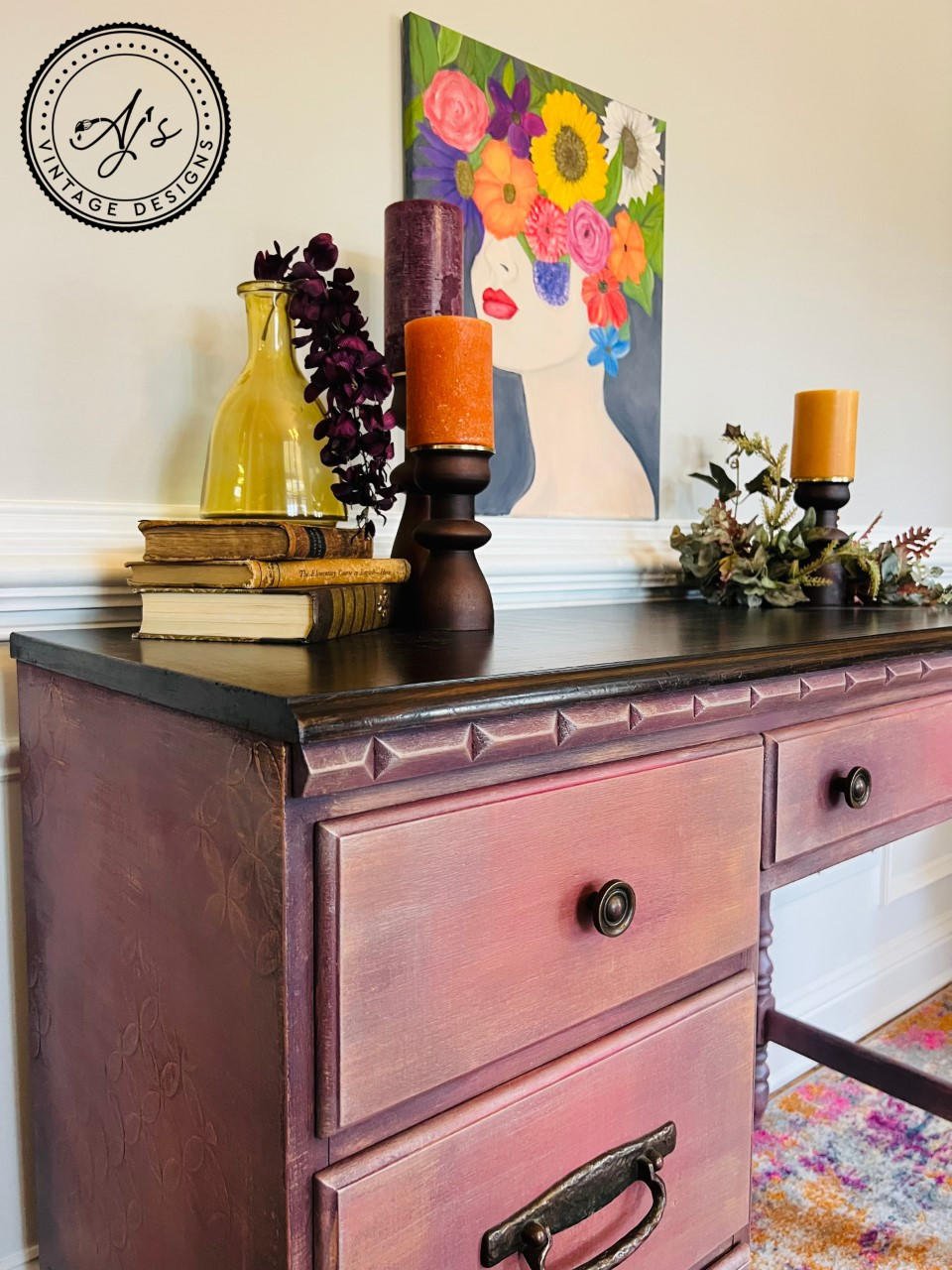 Elderberry Terra Clay Paint provides endless possibilities for your imagination and can be used on wood, canvas, tile, fabric and more! Pink and purple clay painted desk makeover with a dark stained top. Staged with a stack of 3 books with a yellow glass vase containing roses, a large candle stick with a purple candle, 2 candlesticks with orange candles and faux greenery. Large canvas painting on the wall over the desk of a woman with a colorful flower bouquet for hair.