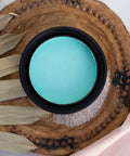 Lani's Lagoon Terra Clay Paint provides endless possibilities for your imagination and can be used on wood, canvas, tile, fabric and more!
Top view of an opened container of the Lani's Lagoon Terra Clay Paint  staged on a round wooden board, a light pink linen and a fan leaf around the piece.