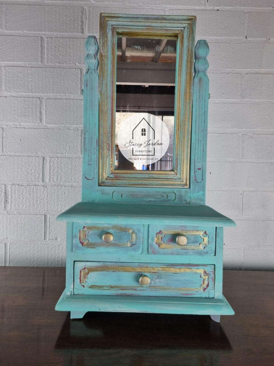 Lani's Lagoon Terra Clay Paint provides endless possibilities for your imagination and can be used on wood, canvas, tile, fabric and more!
A vanity painted in Lani's Lagoon Terra Clay Paint  with gold around the framing of the drawers and mirror. Staged on dark brown floor and white brick background.