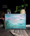 Lani's Lagoon Terra Clay Paint provides endless possibilities for your imagination and can be used on wood, canvas, tile, fabric and more!
A dresser painted in an abstract sunset look with a blend of all the colors in the Terra Clay Paint line including Lani's Lagoon Terra Clay Paint. Staged with greenery, vase and a vintage camera with a woven basket with a faux houseplant in on the opposite side of the dresser top.