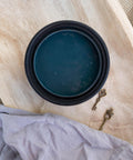 London Blue Terra Clay Paint provides endless possibilities for your imagination and can be used on wood, canvas, tile, fabric and more! 
Top view of an opened container of London Blue Terra Clay Paint staged on top of a light wooden board, greenery and a gray cloth around the piece.