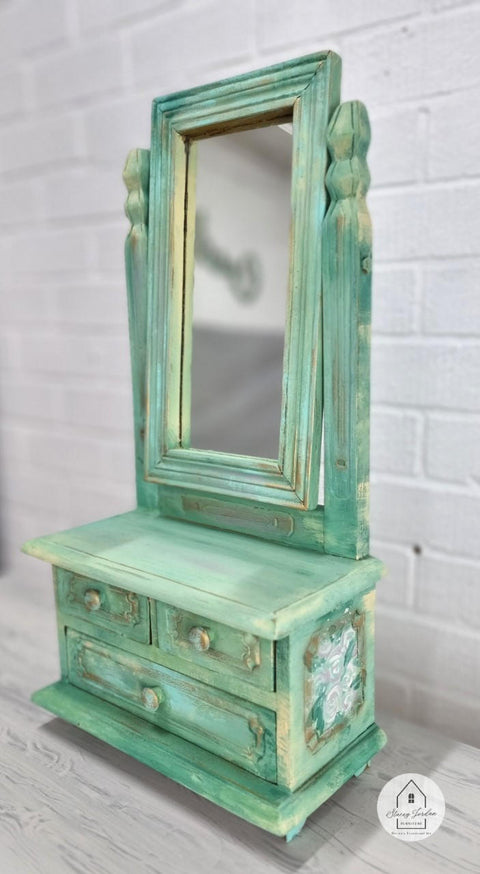 Malachite Terra Clay Paint provides endless possibilities for your imagination and can be used on wood, canvas, tile, fabric and more!
Vanity with rotatable mirror painted in Malachite Terra Clay Paint and with a blend of yellow with brown notes to give it a blended tie dye look. Staged on white wood floors and white brick wall.