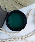 Malachite Terra Clay Paint provides endless possibilities for your imagination and can be used on wood, canvas, tile, fabric and more!
Open container of Malachite Terra Clay Paint staged with white wooden beads, a black book and Large dry leaf around container.