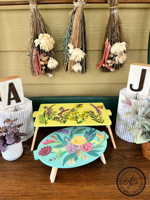 Pistachio Terra Clay Paint provides endless possibilities for your imagination and can be used on wood, canvas, tile, fabric and more! Wood trays clay painted in a yellow and green with painted flowers and rub on transfers. Staged next to a wood a and j block and potted plant.