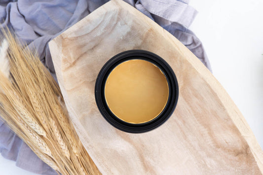 Wheat Terra Clay Paint provides endless possibilities for your imagination and can be used on wood, canvas, tile, fabric and more!  Top view of an open jar of Wheat Terra Clay paint sitting inside a wooden bowl with wheat to the side and a lavender piece of fabric underneath, all on a white table.