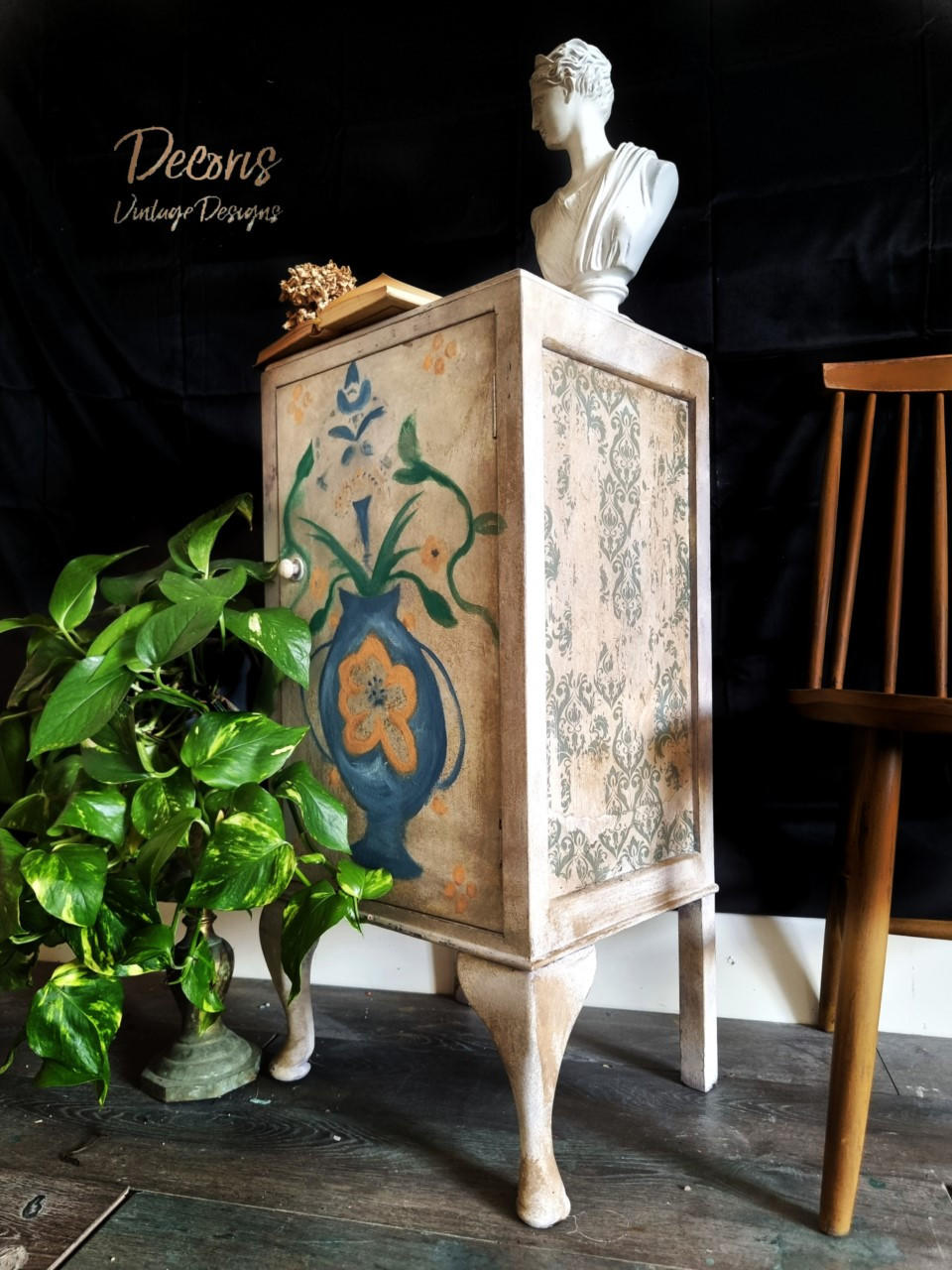 Terra Clay Paint Wheat Terra Clay Paint.  Wheat chalk painted cabinet with a blue, yellow and green vase hand painted on the front of it.  A pattern is stenciled to the side with gold paint.  The cabinet stands on a brown wooden floor with a black background.  A brown chalk painted chair sits to the right of the cabinet and a green plant sits to the left.
