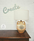 Wheat Terra Clay Paint provides endless possibilities for your imagination and can be used on wood, canvas, tile, fabric and more!  A wheat chalk painted ceramic lamp with a WoodUbend embellishment on the front and a white lampshade.  Sitting on top of a brown wooden table, in front of a white brick wall.  A sign that reads "Create" is hanging on the wall.