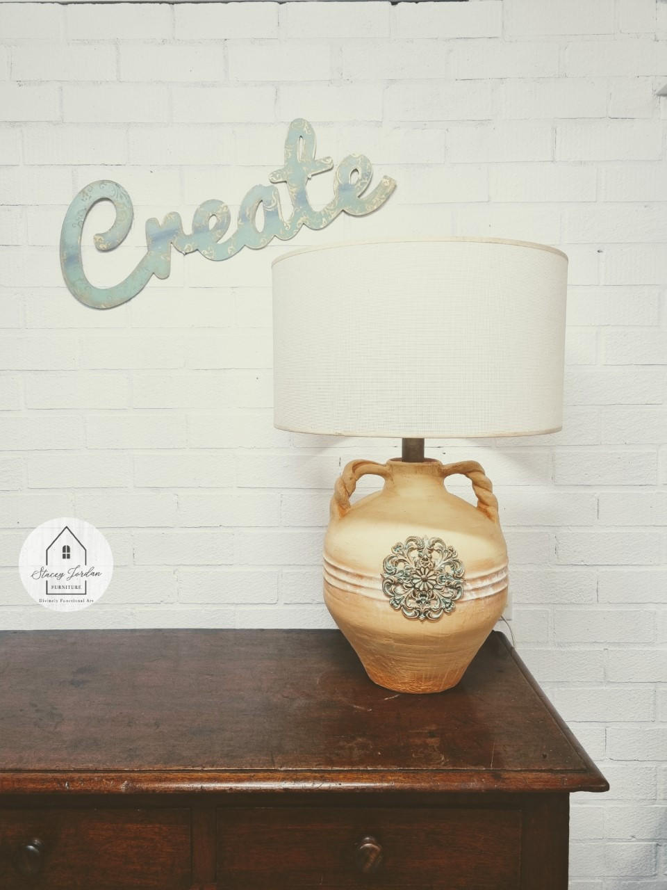 Wheat Terra Clay Paint provides endless possibilities for your imagination and can be used on wood, canvas, tile, fabric and more!  A wheat chalk painted ceramic lamp with a WoodUbend embellishment on the front and a white lampshade.  Sitting on top of a brown wooden table, in front of a white brick wall.  A sign that reads "Create" is hanging on the wall.