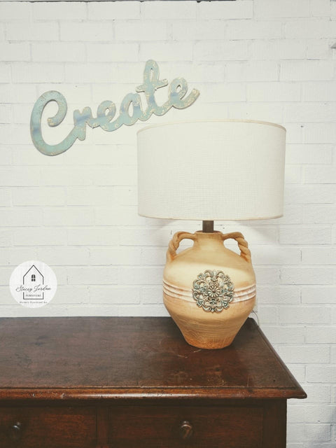 Wheat Terra Clay Paint provides endless possibilities for your imagination and can be used on wood, canvas, tile, fabric and more!  A wheat chalk painted ceramic lamp with a WoodUbend embellishment on the front and a white lampshade.  Sitting on top of a brown wooden table, in front of a white brick wall.  A sign that reads "Create" is hanging on the wall.