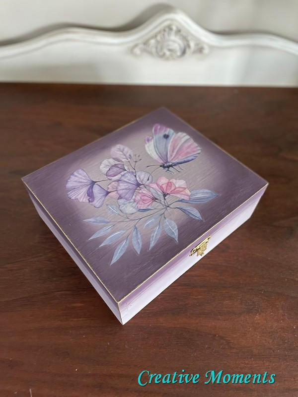 Wisteria Mist Terra Clay Paint provides endless possibilities for your imagination and can be used on wood, canvas, tile, fabric and more!  A small wooden box that has been chalk painted with purple and Wisteria Mist Terra Clay Paint.  The Translucent Garden Transfer is applied to the center of the top of the box, and the clasp is gold.  The box sits on a brown wooden table, in front of a white wall.