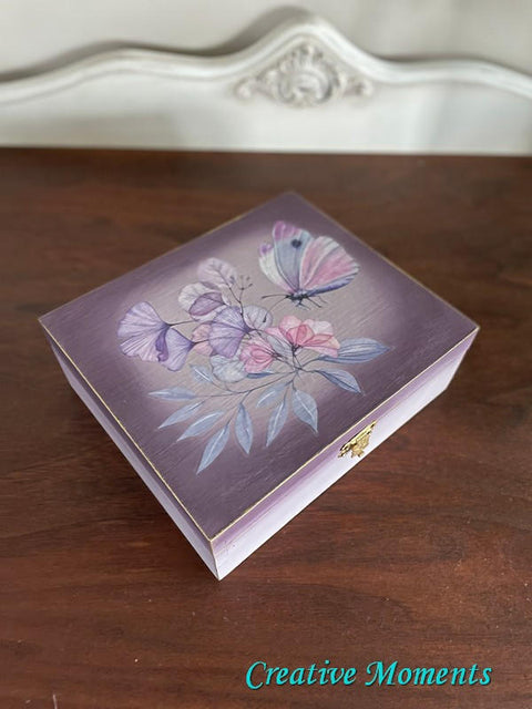 Wisteria Mist Terra Clay Paint provides endless possibilities for your imagination and can be used on wood, canvas, tile, fabric and more!  A small wooden box that has been chalk painted with purple and Wisteria Mist Terra Clay Paint.  The Translucent Garden Transfer is applied to the center of the top of the box, and the clasp is gold.  The box sits on a brown wooden table, in front of a white wall.