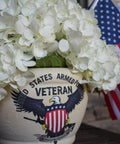 A flower pot with "United States Armed Forces Veteran" on the front, decorated with a U.S. flag emblem and an eagle, holding lush white hydrangeas, with a small American flag visible in the background.