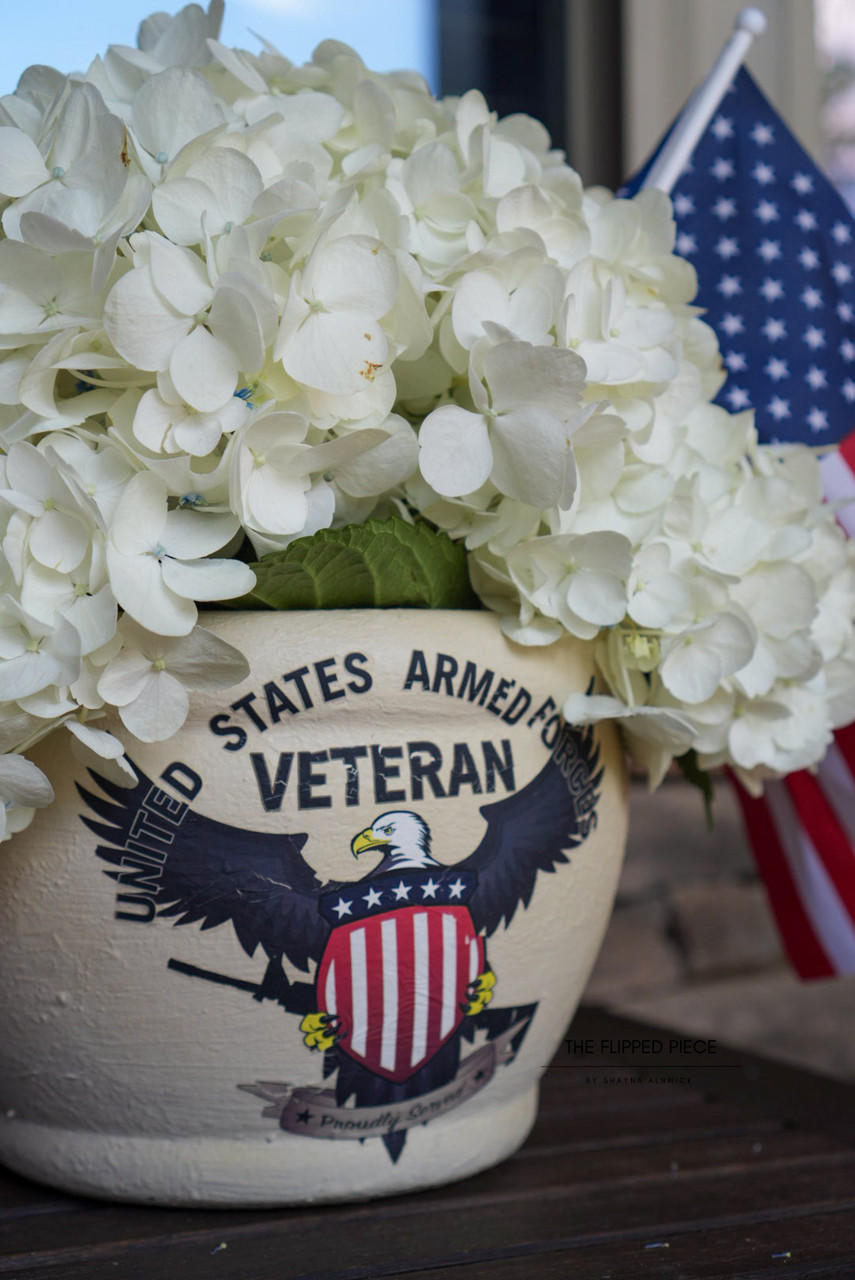 A flower pot with "United States Armed Forces Veteran" on the front, decorated with a U.S. flag emblem and an eagle, holding lush white hydrangeas, with a small American flag visible in the background.