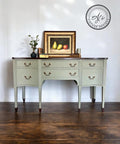 A vintage-style sideboard with light green drawers and dark wood accents stands against a white wall. On it rests a framed painting of pears and an apple, accompanied by a small plant and decorative items. A circular watermark is on the wall above.