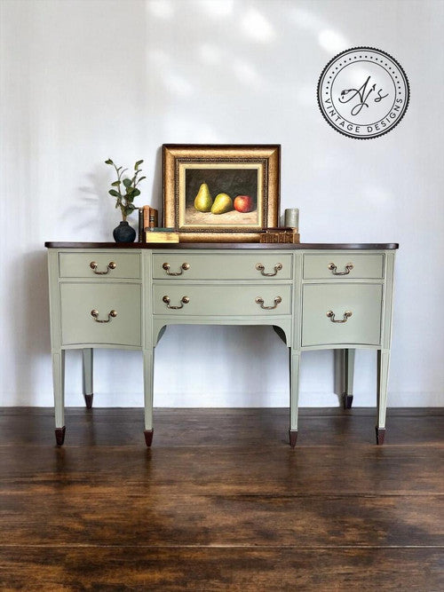 A vintage-style sideboard with light green drawers and dark wood accents stands against a white wall. On it rests a framed painting of pears and an apple, accompanied by a small plant and decorative items. A circular watermark is on the wall above.