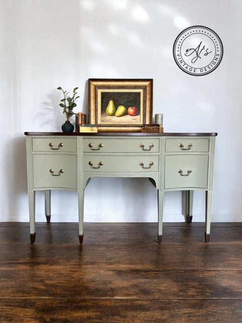A vintage-style sideboard with light green drawers and dark wood accents stands against a white wall. On it rests a framed painting of pears and an apple, accompanied by a small plant and decorative items. A circular watermark is on the wall above.