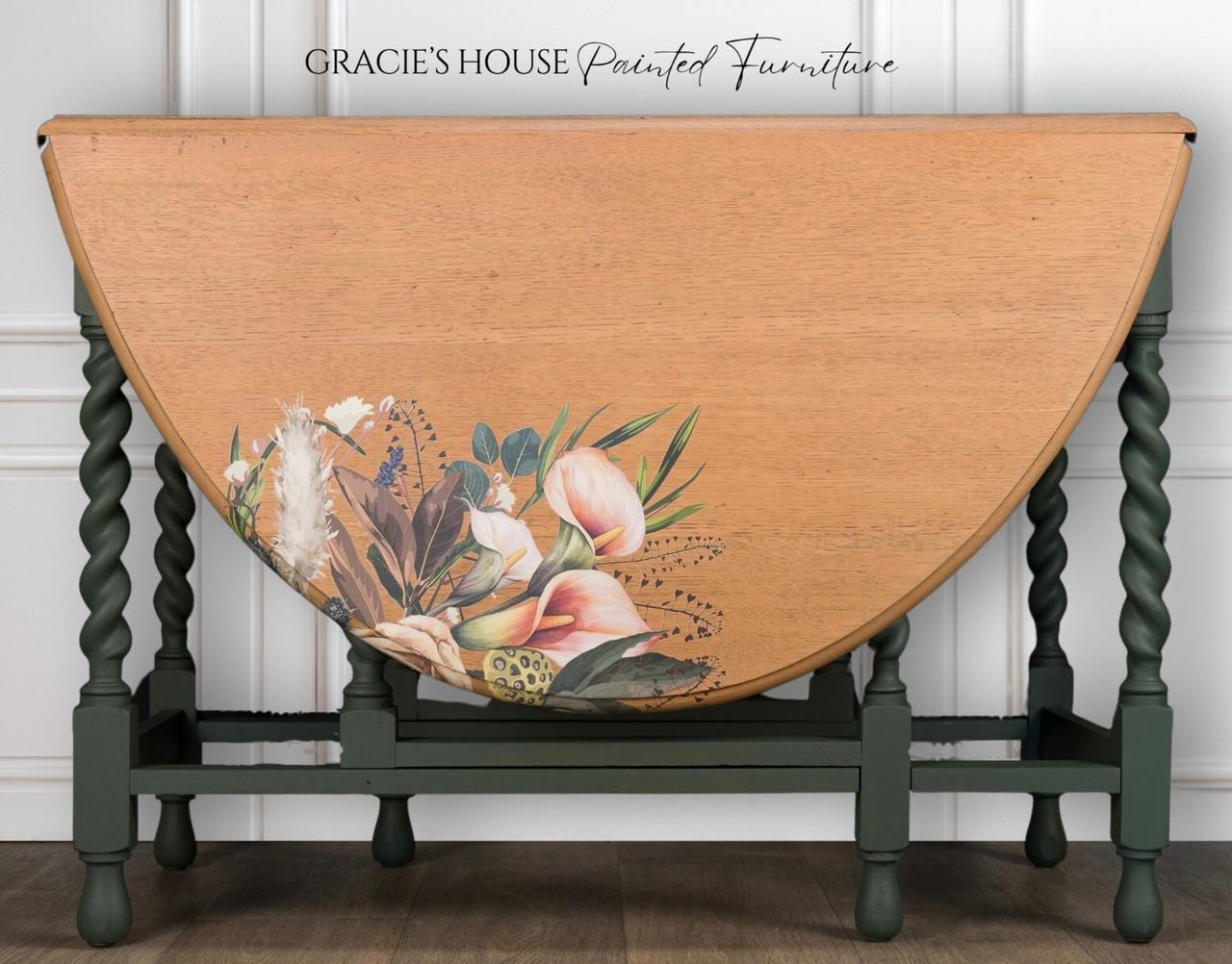 A vintage wooden table with turned green legs features a drop-leaf design. The tabletop displays a floral design with various flowers and leaves. The background has white paneled walls with "Gracie's House Painted Furniture" written above.