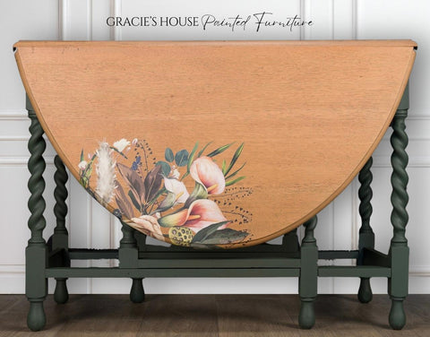 A vintage wooden table with turned green legs features a drop-leaf design. The tabletop displays a floral design with various flowers and leaves. The background has white paneled walls with "Gracie's House Painted Furniture" written above.