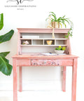 A lovely pink and white blended desk with the Breast Cancer awareness decoupage paper on the front drawer.  Plants in pots are staged on the piece as well as a pen and notebook.