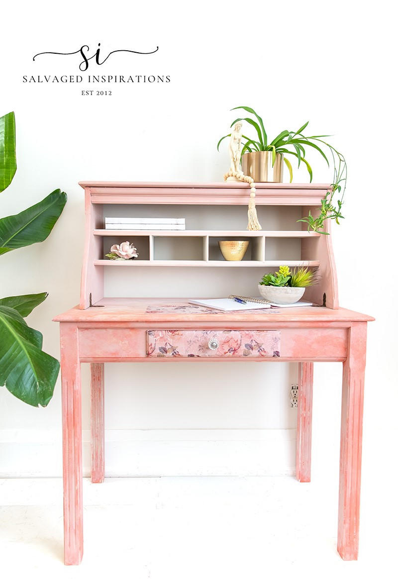 A lovely pink and white blended desk with the Breast Cancer awareness decoupage paper on the front drawer.  Plants in pots are staged on the piece as well as a pen and notebook.