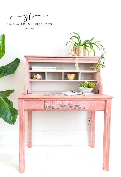 A lovely pink and white blended desk with the Breast Cancer awareness decoupage paper on the front drawer.  Plants in pots are staged on the piece as well as a pen and notebook.