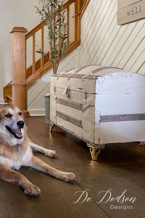 A chest that has been chalk painted in white and staged in front of a staircase.  A dog is sitting to the side of it.