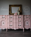 A pink dresser that is blended with some white chalk paint in the middle near the handles creating a ombre effect.  staged with an empty gold picture frame and candle light and some flowers.
