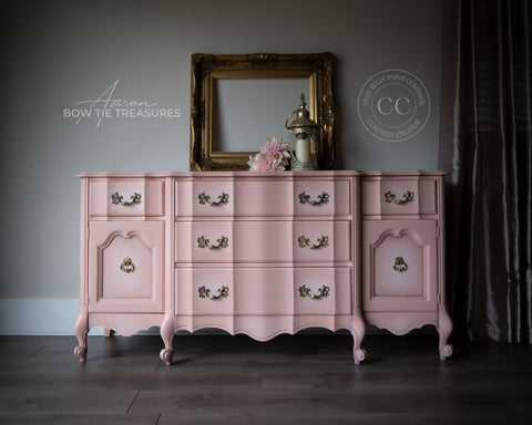 A pink dresser that is blended with some white chalk paint in the middle near the handles creating a ombre effect.  staged with an empty gold picture frame and candle light and some flowers.